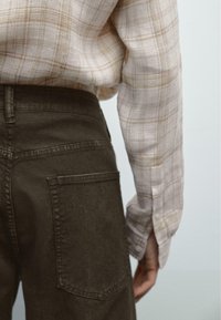 Světle kostkovaná košile s béžovými a hnědými pruhy, spojená s tmavě zelenými kalhotami s kapsou na zadní straně a rovnými nohavicemi.