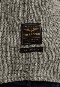 Siva tkanina z vzorcem, ki vključuje znak "PME Legend" v zlatem tisku in grafiko peruti. Pod njim je črna oznaka z napisom "Typ N°: C-46."