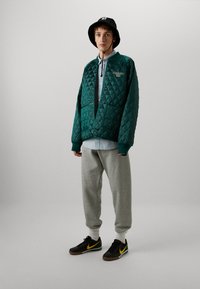 Chaqueta verde acolchada con cierre de botón, camiseta gris debajo, pantalones de chándal grises y zapatillas negras con detalles amarillos. Sombrero tipo cubo negro.