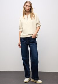 Suéter beige claro con cuello de media cremallera, combinado con vaqueros de pierna ancha de color azul oscuro y zapatillas blancas, de pie frente a un fondo neutro.