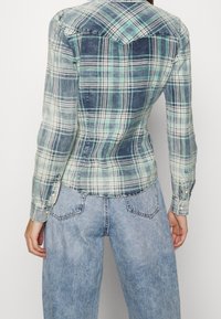 Chemise à carreaux aux nuances de bleu, vert et blanc. Manches longues avec poignets boutonnés. Coupe ajustée avec ourlet incurvé et détail de pièce en dos.