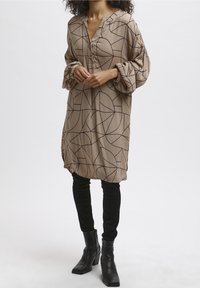 Robe beige à motif avec un décolleté en V et de longues manches froncées ; présente des lignes géométriques noires ; assortie à un pantalon noir ajusté et des bottes à chevilles.