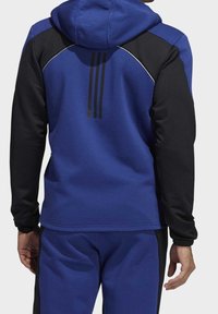 Sweat à capuche bleu avec manches noires, tissu texturé, trois bandes noires dans le dos et surpiqûres blanches le long des coutures.