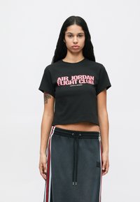 Γυναίκα με μακριά σκοτεινά μαλλιά, φορώντας μαύρο κοντό μπλουζάκι με ροζ κείμενο "Air Jordan Flight Club" και σκούρες φόρμες με κόκκινες και λευκές ρίγες στο πλάι.