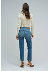 Mujer de pie de espaldas, vistiendo una blusa de manga larga color crema, jeans de tiro alto azules y sandalias de tacón marrones, sobre un fondo gris.
