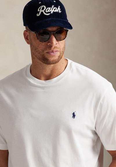 Bílé bavlněné tričko s malým námořnickým logem na hrudi, doplněné o námořnickou manšestrovou čepici s nápisem „Ralph“ v bílém písmu a hnědé sluneční brýle.