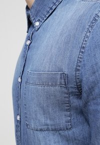 Camisa de mezclilla azul clara con cuello abotonado, bolsillo frontal y una tela sutilmente texturizada con un diseño de corte clásico.