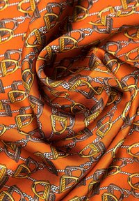Foulard di seta con sfondo arancione, caratterizzato da un motivo ripetuto di staffe marroni e gialle intrecciate con corde bianche. Texture liscia.