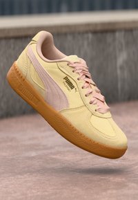 Puma sneakers with a light yellow suede upper, pink accents, and a textured orange rubber sole, featuring a logo on the side and pink laces.