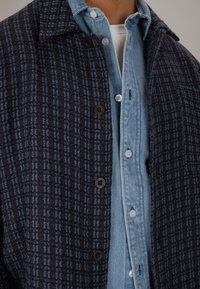 Teksturert mørkeblå yttertøy med rutet mønster, med knappestolpe foran og en lyseblå denimskjorte under.