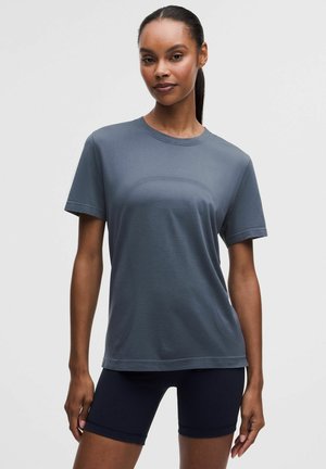 Frau steht, trägt ein kurzärmeliges graues strukturiertes Shirt und schwarze Radfahrshorts vor einem einfarbigen hellen Hintergrund.