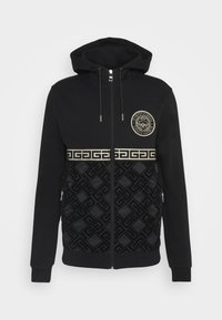 Černá mikina na zip s kapucí, zdobená emblémem lva, zlatým vodorovným pruhem a texturovaným geometrickým vzorem ve spodní části.
