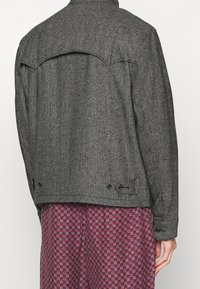 Grå herringbone-mönstrad jacka med bakre yoke, sidofickor och knapppynt. Slät textur, figurnära design och tjockt tyg.