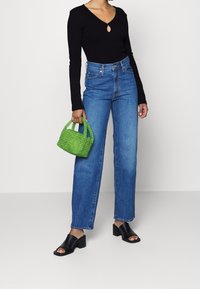 Schwarzes langärmliges Oberteil mit Schlitzdesign, blaue hochtaillierte Jeans, grüne geflochtene Handtasche und schwarze offene Hi-Heels.