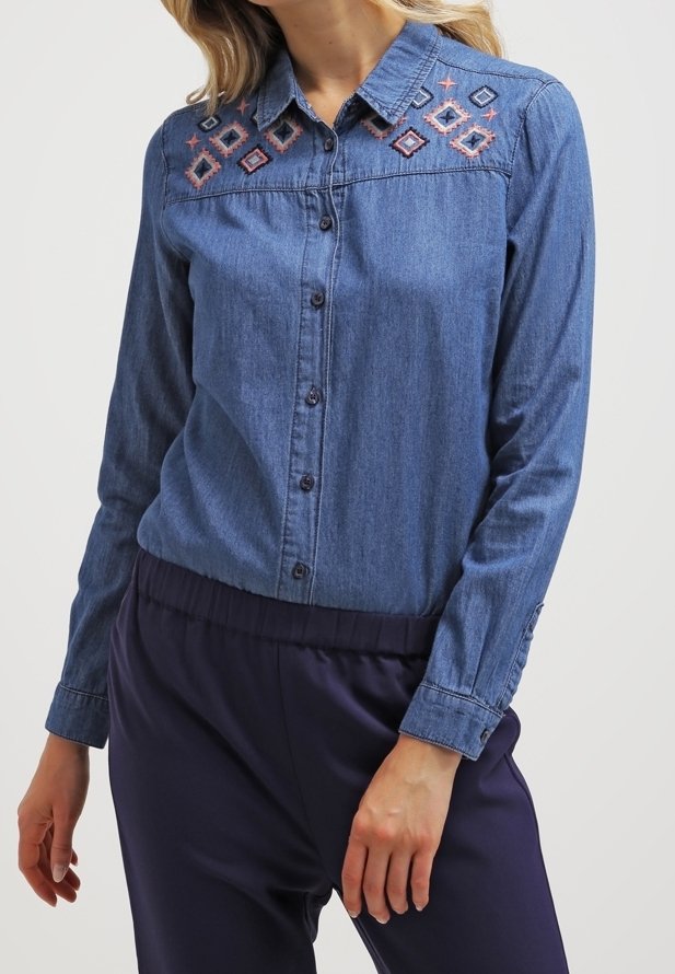 Femme portant une chemise en jean bleue à boutons avec des motifs géométriques brodés sur la partie supérieure de la poitrine et un pantalon bleu foncé.