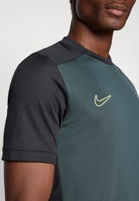 Tričko Nike s krátkym rukávom má tmavozelený predný diel a čierne rukávy, je vyrobené z hladkého materiálu s jemným leskom a s logom na hrudi.