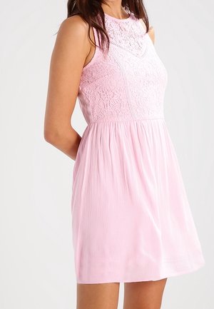 Woman wearing sleeveless light pink dress with lace bodice and gathered skirt, standing with hands behind her back.