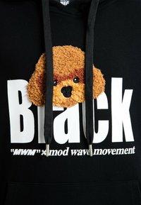 Sudadera negra con gran texto en blanco, una cara de perro texturizada en marrón y canela, y cordones negros con puntas plateadas.