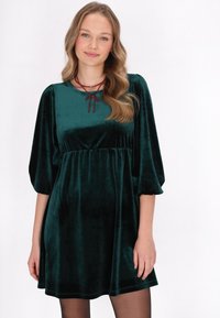 Rochie din catifea verde smarald, cu decolteu rotund, mâneci bufante și talie empire. Are un fundiță decorativă la decolteu.