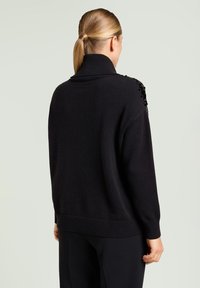 Suéter negro de gran tamaño con cuello alto acanalado, tela texturizada y detalles adornados en los hombros. Lleva pantalones oscuros.