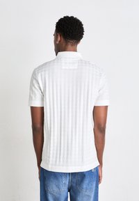 Hvit kortermet polo med strukturmønster, krage og bakparti-detalj, kombinert med blå jeans.