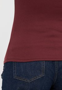 Bordová upnutá tílko s ribovanou strukturou, umístěné nad tmavě modrými džíny. Viditelné švy a boční rozparky.