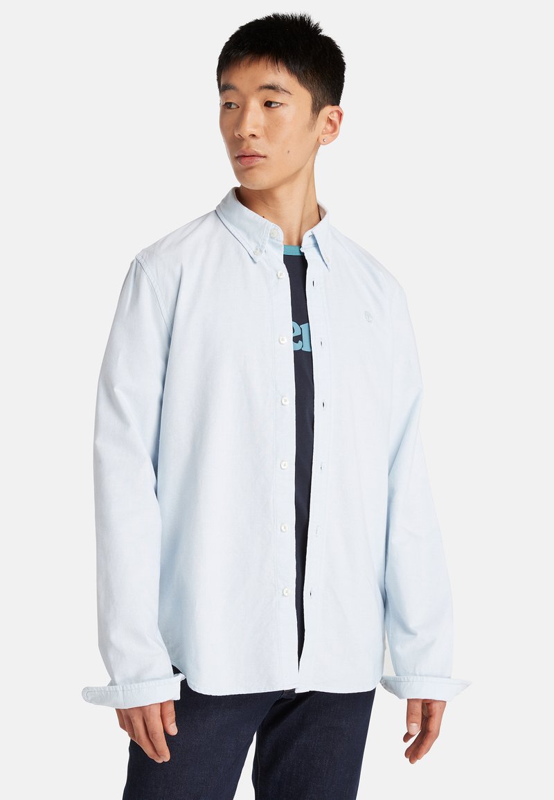 Lichtblauw, knoopsluiting overhemd met lange mouwen en een kraag, gedragen over een donkermon tshirt. Heldere stof met een gladde textuur.