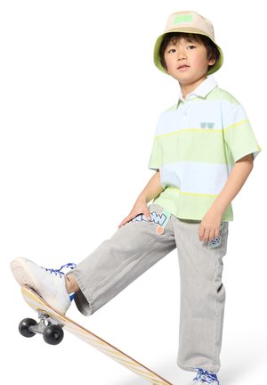 Polo rayé vert clair et bleu, pantalon gris avec des patchs colorés, baskets blanches avec lacets bleus, chapeau seau beige, sur un skateboard.