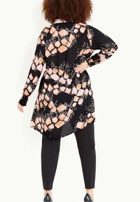 Blouse longue noire présentant un motif de formes pêche et blanches avec un ourlet arrondi, associée à un pantalon noir slim et des chaussures fermées.