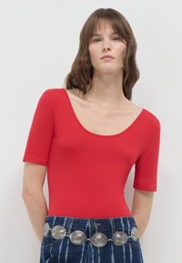 Donna che indossa una maglietta rossa con scollo a barca e jeans in denim blu a vita alta, con una cintura a medaglione d'argento, in piedi contro uno sfondo chiaro e semplice.