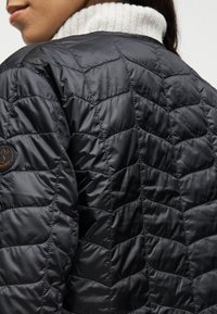 Κοντινό πλάνο ατόμου που φοράει μαύρο μπουφάν με ραφές σε σχήμα V και λευκή ζιβανγκιέρ πλεκτή στο λαιμό, ορατή στον γιακά.