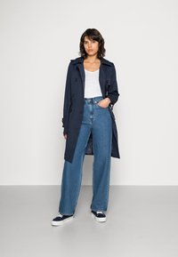 Jūrinis trench paltas su dvigubais mygtukais, dideliais juodais mygtukais, dėvimas ant baltos marškinios ir aukštu liemeniu mėlynų džinsų. Baltos sportbačiai.