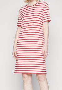 Femme portant une robe blanche mi-longue avec des rayures horizontales rouges et des manches courtes, debout devant un fond uni.