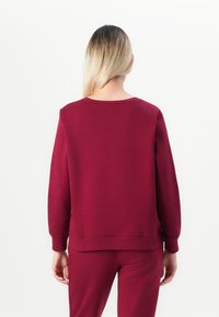 Woman with long blonde hair wearing a plain burgundy sweatshirt and matching pants, standing facing away against a white background.