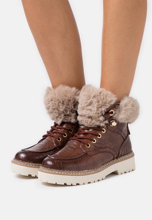 Bottes de cheville en faux cuir marron avec une finition texturée, des revers en faux fourrure beiges, des œillets métalliques et une semelle en caoutchouc blanche épaisse.