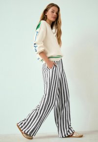 Hanorac crem cu accente verzi și text albastru pe mâneci, asortat cu pantaloni evazați cu dungi negre și albe și pantofi bej.