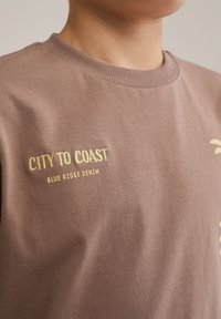 Rudas medvilninis marškinėliai su auksiniu siuvinėjimu, kuriame užrašyta "CITY TO COAST" ir "BLUE RIDGE DENIM" priekyje; su įribbtu apvaliu kaklu.