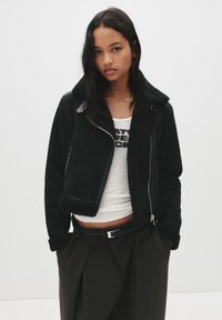 Giacca in shearling nera con chiusura a zip, ampio colletto e foderato con una texture, indossata sopra un top bianco aderente e pantaloni larghi scuri.