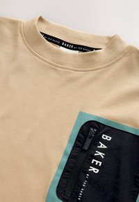 T-shirt beige à col rond avec une poche zippée bleu sarcelle et noire sur la poitrine, marqué « Baker by Ted Baker » sur la poche et le col.
