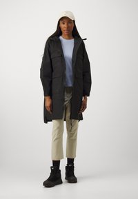 Parka noire longue avec plusieurs poches, fermeture éclair, capuche réglable, associée à un t-shirt bleu clair et un pantalon beige, bottes noires.