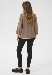 Femme aux longs cheveux ondulés portant un pull marron ample, un pantalon noir slim et des mocassins compensés noirs, debout de dos sur un fond blanc.