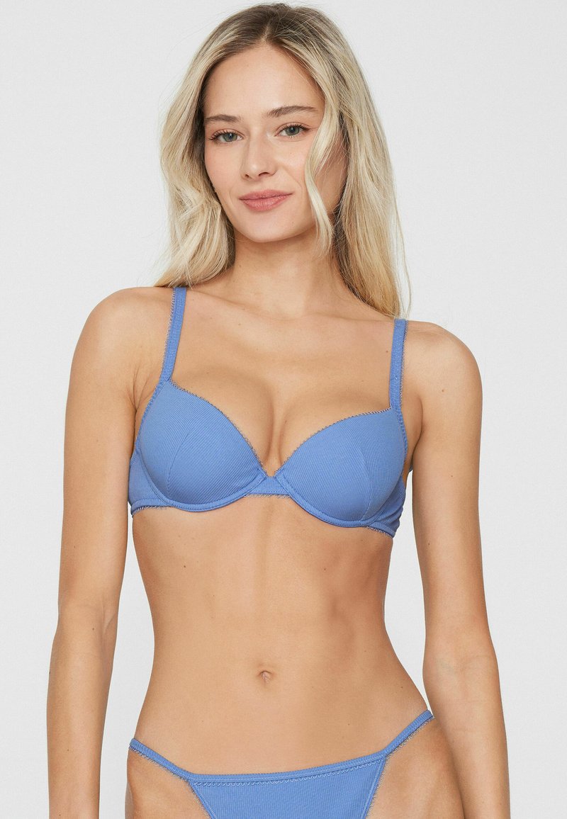 Mujer con cabello rubio largo vistiendo un conjunto a juego de sujetador acanalado azul claro y ropa interior estilo bikini, de pie contra un fondo claro y liso.