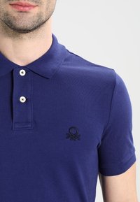 Polo a maniche corte di colore blu scuro, realizzato in tessuto strutturato. Presenta un logo ricamato nero sul petto sinistro e una pattina con due bottoni.
