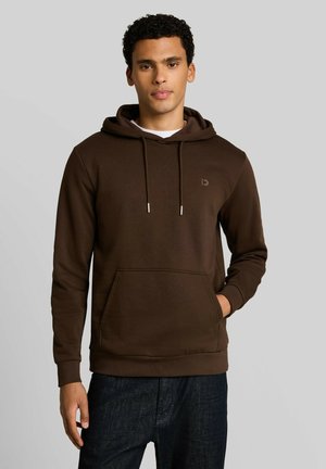 Junger Mann trägt einen dunkelbraunen Kapuzenpullover mit Kordeln und einer Fronttasche, steht vor einem schlichten hellgrauen Hintergrund.