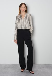 Chemise à boutons avec des rayures ondulées noires et beiges, associée à un pantalon noir ajusté. Le modèle se tient devant un fond neutre.
