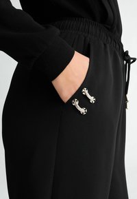 Ręka w kieszeni czarnych spodni z elastycznym pasem i srebrnymi ozdobami z kamieniami przy krawędzi kieszeni.