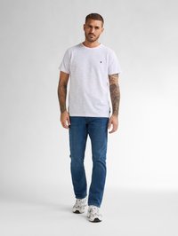 Tricou alb cu dungi colorate și guler rotund, asociat cu blugi blue. Bărbatul poartă adidași albi. Sunt vizibile tatuaje pe brațele lui.