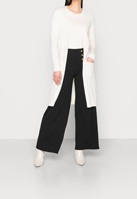 Dlouhý krémový cardigan s texturovanou tkaninou, otevřeným předním dílem a bočními kapsami, v kombinaci s černými kalhotami s širokými nohavicemi, které mají zlaté knoflíkové akcenty.