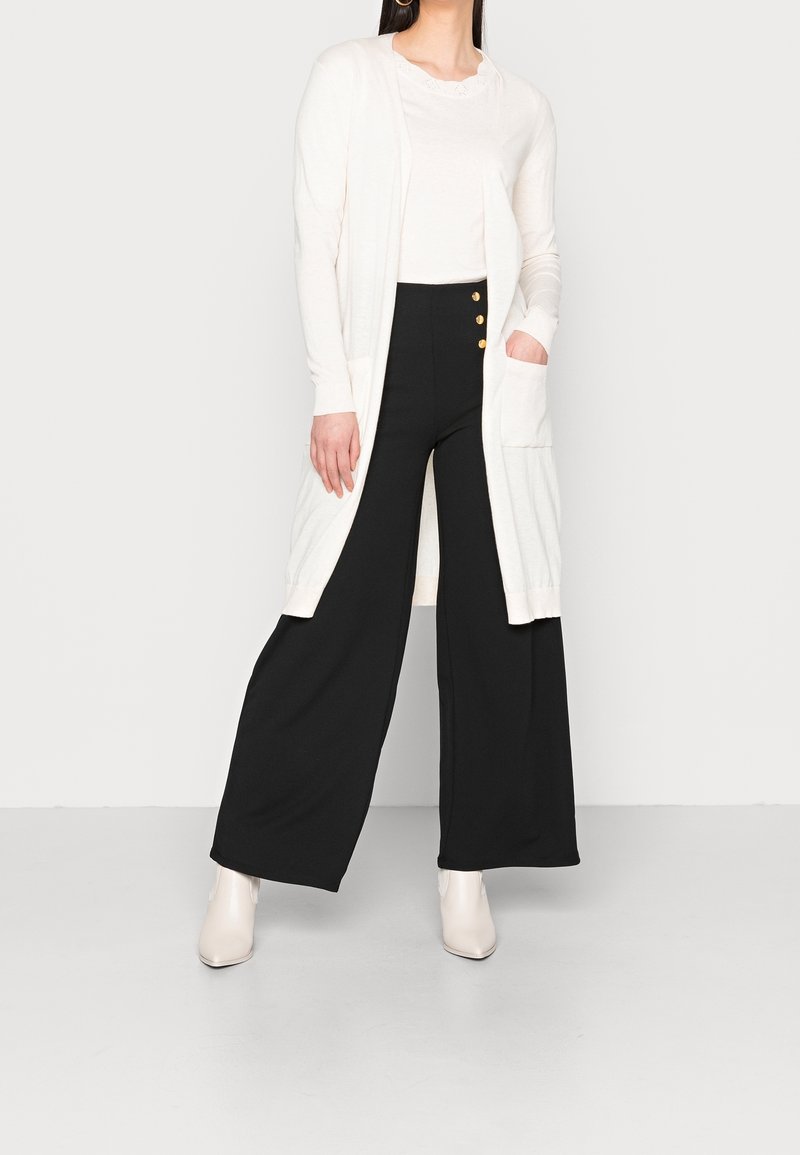 Dlouhý krémový cardigan s texturovanou tkaninou, otevřeným předním dílem a bočními kapsami, v kombinaci s černými kalhotami s širokými nohavicemi, které mají zlaté knoflíkové akcenty.
