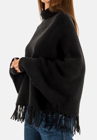 Pull-over en tricot noir côtelé avec un col haut, des manches larges et un ourlet à franges. Le tissu a une apparence douce et texturée.